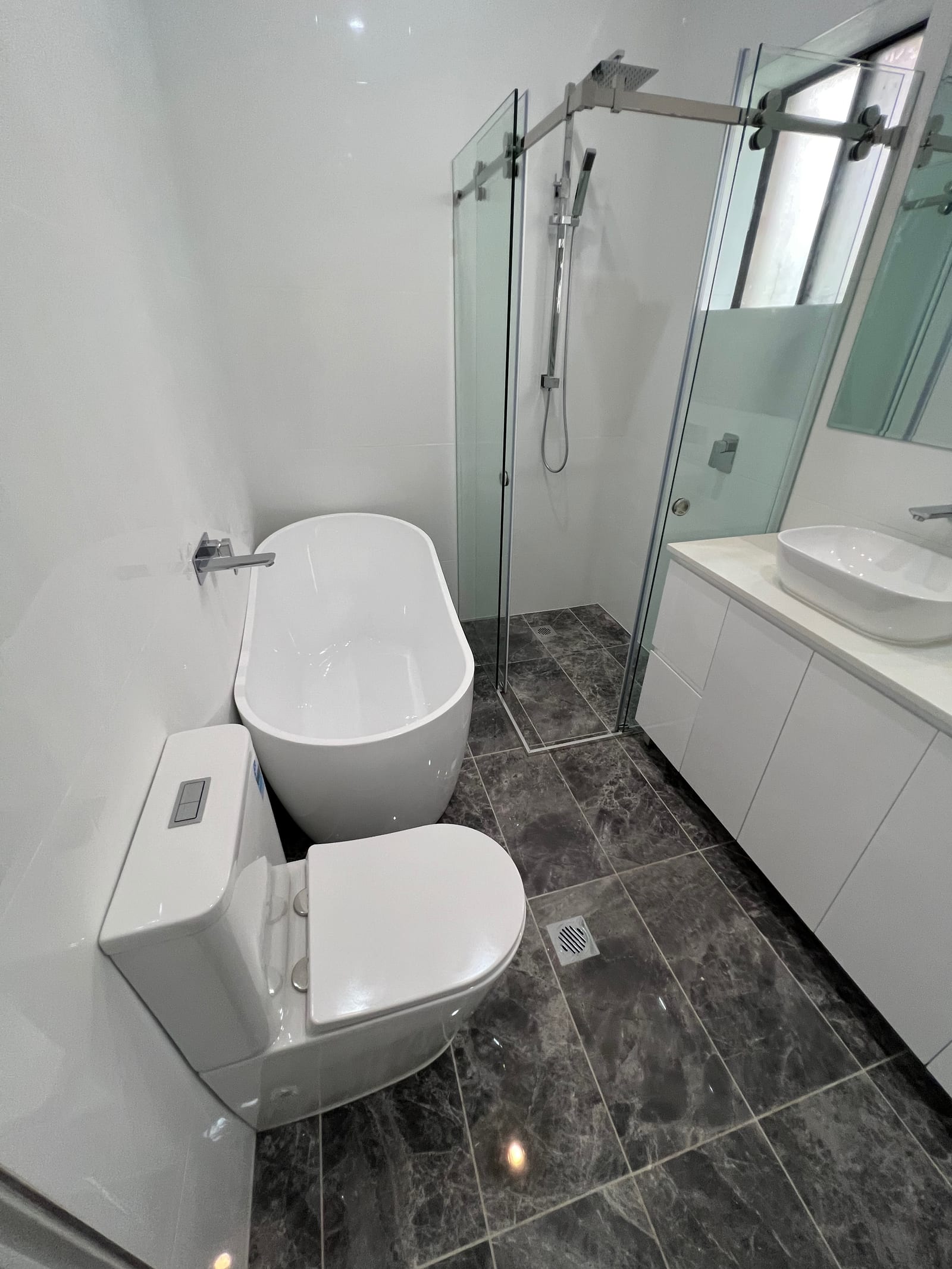 A second finished bathroom — a freestanding bath, a frameless glass corner shower, dark stone-look tiles and a floating vanity