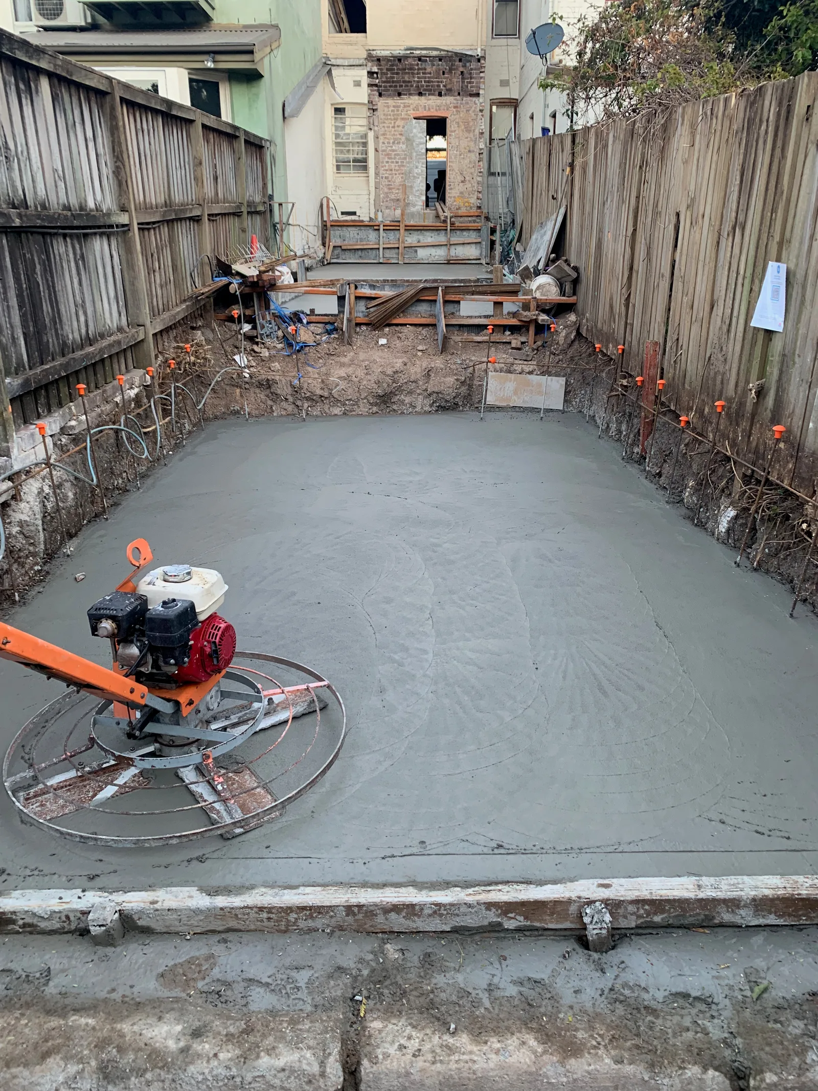 A newly poured and screeded concrete slab for the new construction at the rear of the site, with reinforcement bar exposed at the boundary fence.