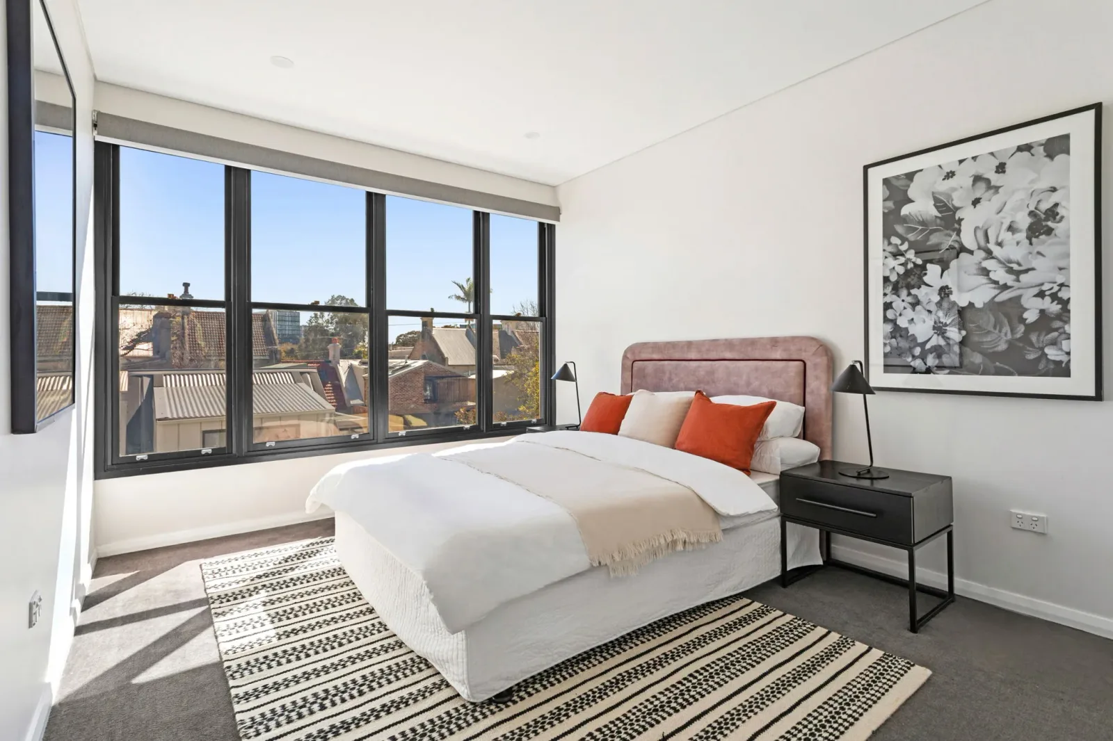 A bedroom with a run of windows looking out over neighbouring rooftops, furnished with an upholstered bedhead and bedside lighting.