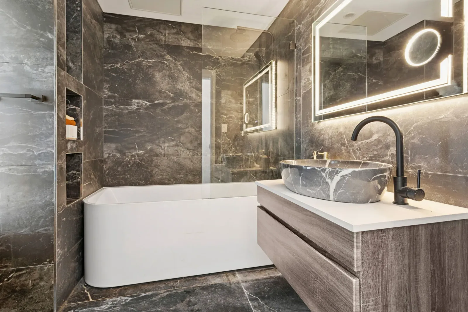 A second bathroom finished in dark stone-look tile, with a freestanding bath, matte-black tapware, an illuminated mirror and a round stone basin on a timber vanity.