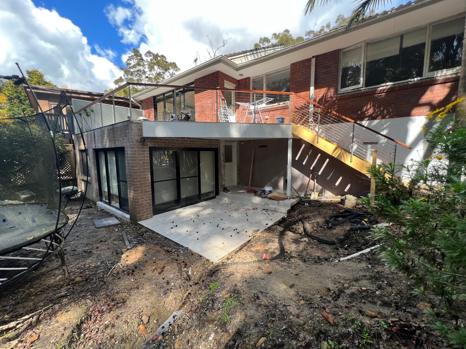 The completed new lower-ground level at the rear of the Lindfield home — a brick-clad wall of sliding glass doors opening onto a paved patio, with a new stainless-steel-balustrade staircase leading up to the existing house above