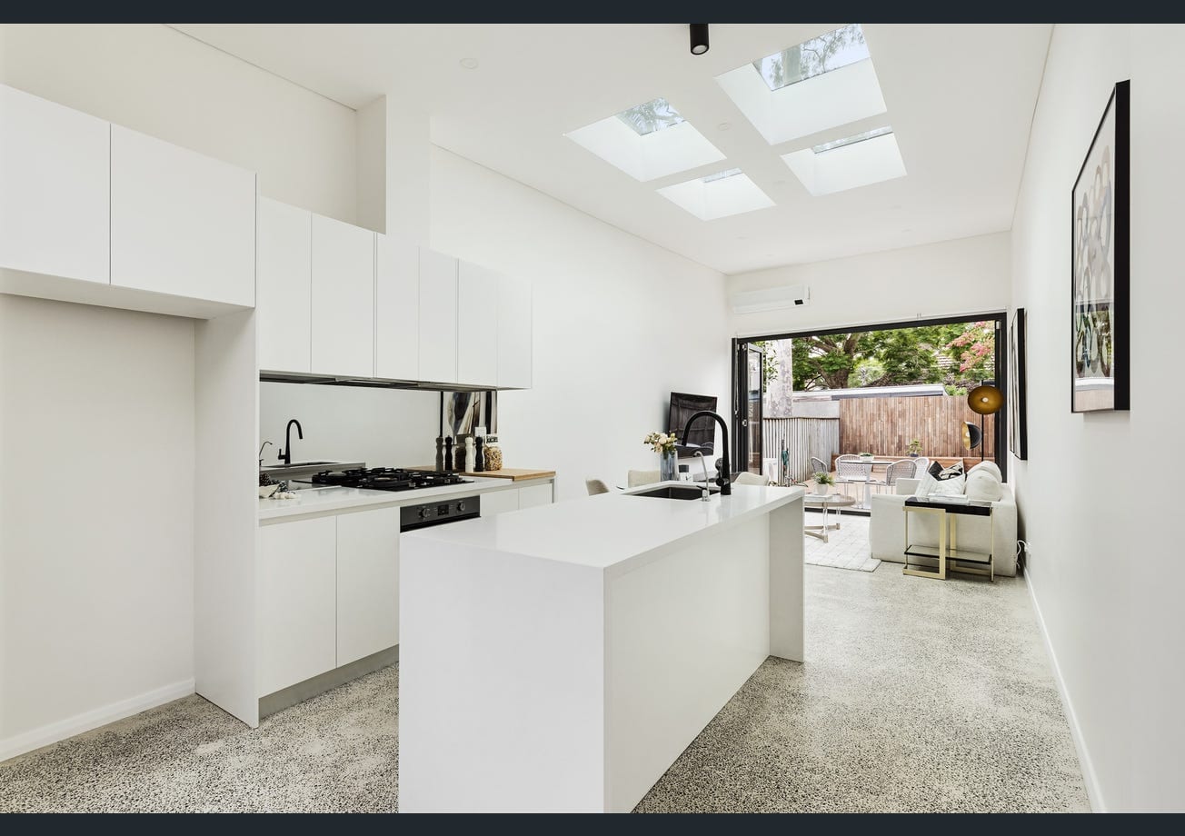 The finished kitchen and open-plan living space — a white island bench, gas cooktop and four skylights overhead, with black-framed glazed doors opening to the rear yard