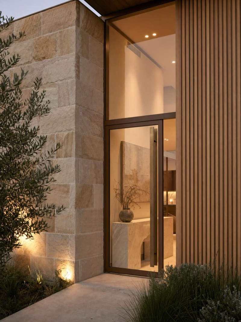 Bespoke new-home entry — honed sandstone, spotted-gum timber battens and full-height glazing at dusk — illustrative concept